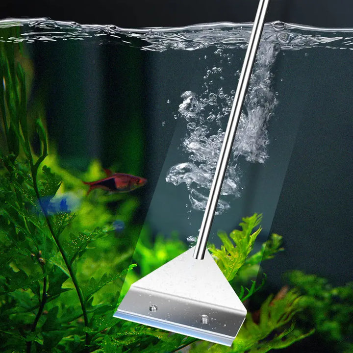 A stainless steel aquarium scraper submerged in water, surrounded by green aquatic plants and bubbles, with a fish swimming nearby.
